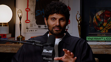 Man with curly hair speaks into a microphone labeled "On Purpose" in a cozy studio with books, awards, a lamp, and wall art in the background, suggesting a podcast or interview setting.