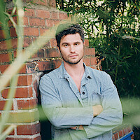 Man in a striped shirt stands with arms crossed by a brick structure, surrounded by green bamboo and foliage, looking directly at the camera in an outdoor garden setting.