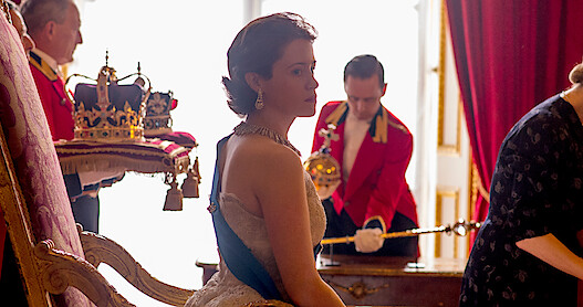 Claire Foy as the Queen in The Crown. She's sitting behind a desk with the royal crown behind her