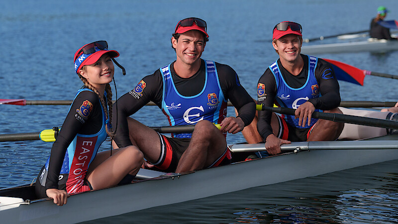 Three rowers wearing uniforms and red caps sit smiling in a racing boat on calm blue water, oars in hand, under clear daylight; outdoor athletic setting, relaxed and cheerful atmosphere.