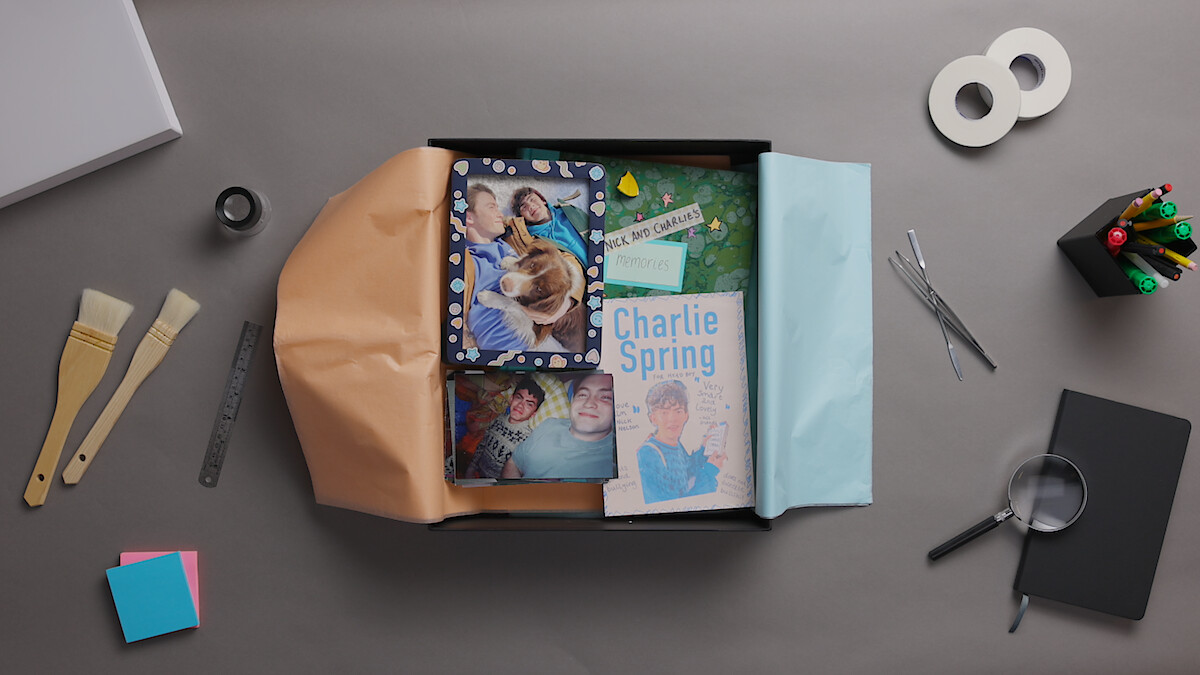 Scrapbook gift box with photos, book titled "Charlie Spring," and keepsakes on a desk. Surrounding items include paintbrushes, ruler, tape, pens, sticky notes, magnifying glass, scissors, and colored paper. Creative workspace setting.