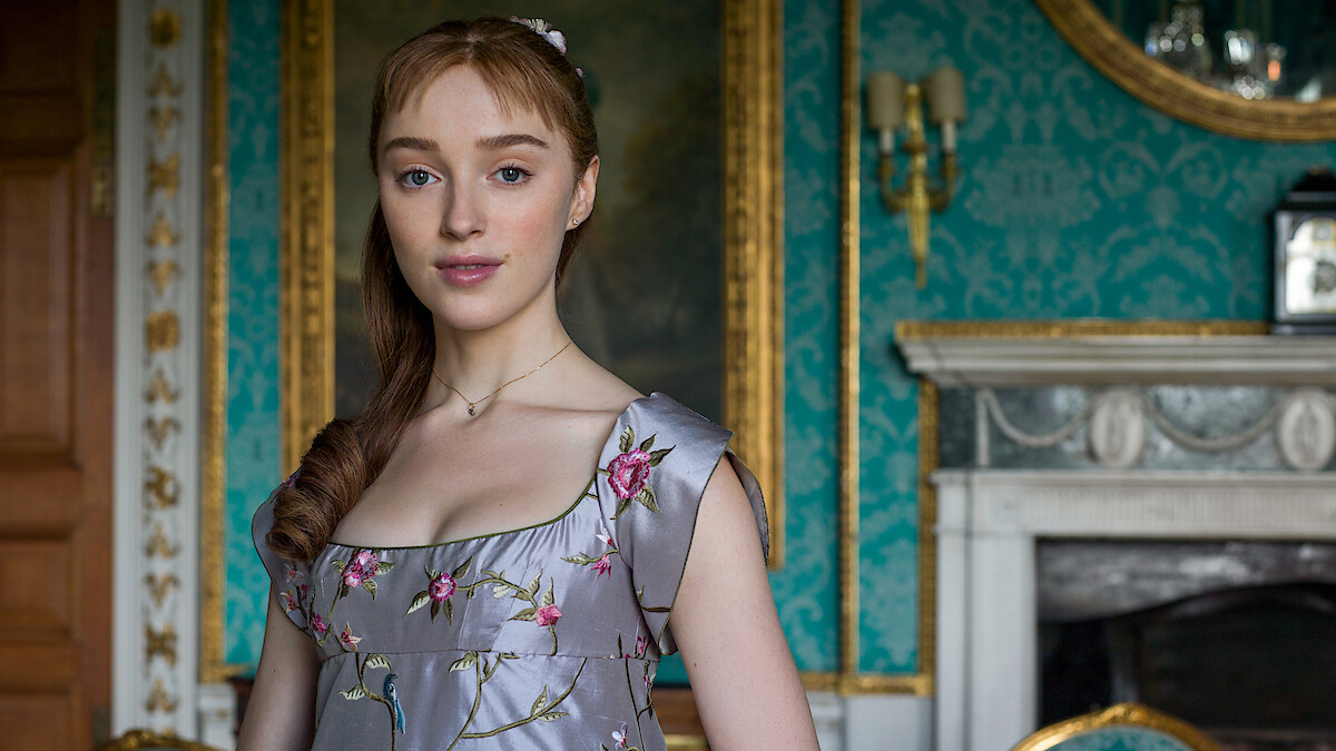 Phoebe Dynevor as Daphne Bridgerton in a lavender, floral embroidered Regency dress stands in an ornate room with teal wallpaper, gold-framed mirror, chandelier, and marble fireplace.