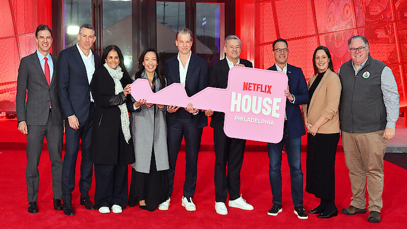 A group of people stand smiling on a red carpet in front of a building, holding a large pink key with "Netflix House Philadelphia" written on it, celebrating an event in a festive, welcoming atmosphere.