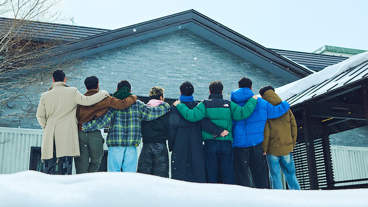 A group of eight people stand arm-in-arm, facing a snow-covered house, dressed warmly in winter clothing. The environment is wintry and serene, conveying friendship and togetherness in a peaceful outdoor setting.