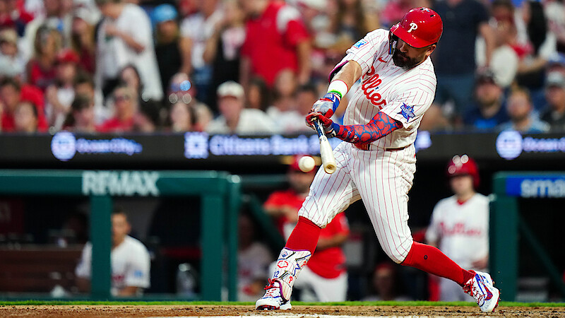 Philadelphia Phillies baseball player in white pinstripe uniform hitting the ball during a game, with a crowd of fans in the stadium background. Daytime, action shot at home plate.