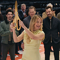 Woman in yellow dress holding large ceremonial scissors, preparing to cut a ribbon at an indoor event with smiling people clapping in the background, suggesting a celebration or grand opening on a basketball court.