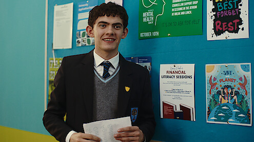 A student in a school uniform stands indoors holding papers, smiling in front of a blue bulletin board with posters about mental health, financial literacy, and environmental awareness.