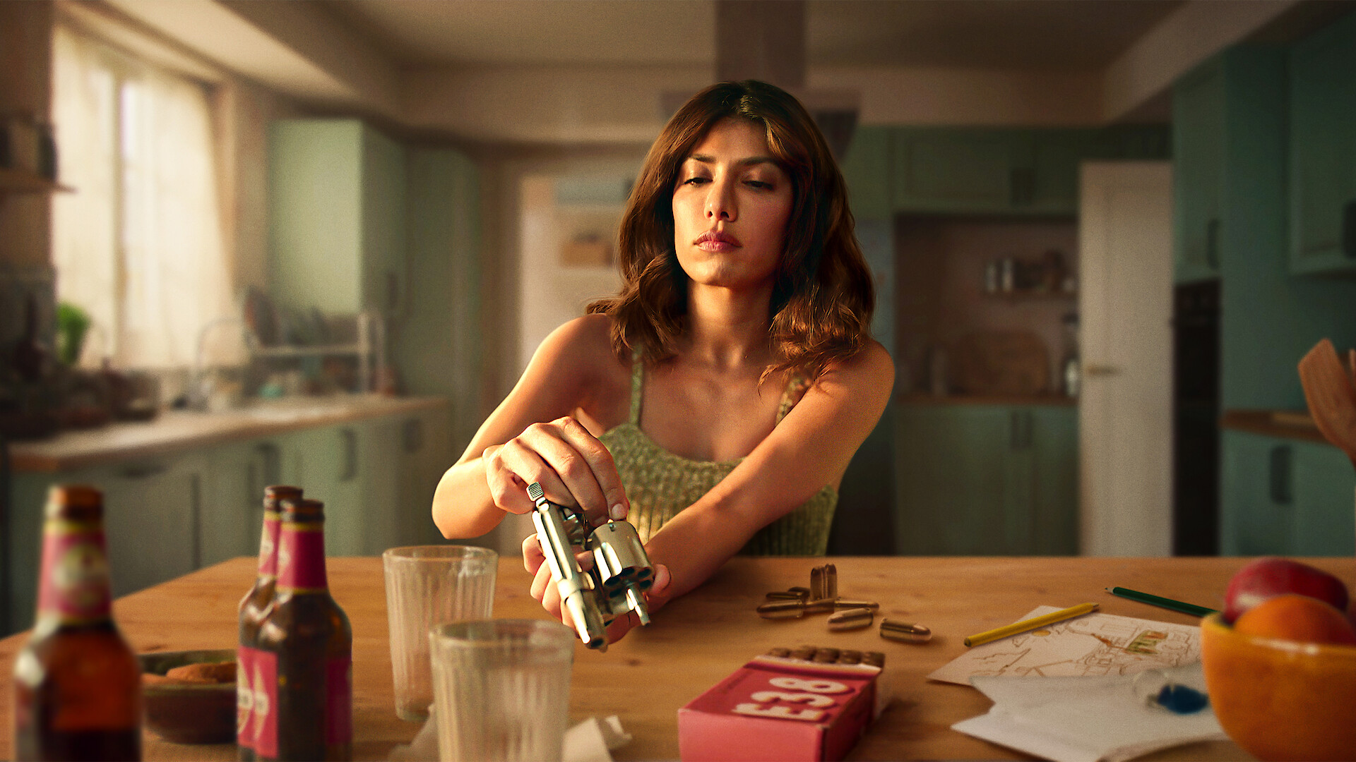 Woman in a kitchen sits at a table with bottles, glasses, and snacks, loading bullets into a revolver, with papers, a red box, and fruit visible, suggesting tension in a domestic setting.