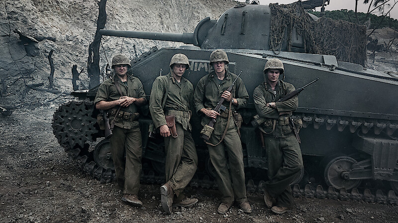 Four soldiers in WWII uniforms stand in front of a tank in a war-torn, smoky environment, suggesting a battlefield setting.