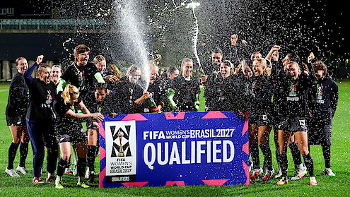 Women's soccer team celebrates at night on a field, spraying champagne and cheering behind a large sign reading "FIFA Women's World Cup Brasil 2027 Qualified." Bright stadium lights and festive atmosphere.