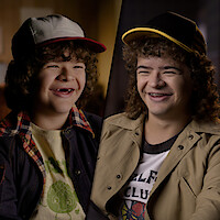 A split-screen image featuring a younger Gaten Matarazzo alongside an older teen version of him, both dressed in casual attire with baseball caps, smiling warmly in softly lit indoor settings.