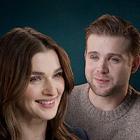 Two people smiling in a close-up portrait with a blurred blue background, one woman with brown hair in the foreground and one man with short hair and a sweater in the background, both appearing friendly and relaxed.