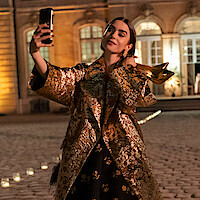 A woman in a luxurious gold-patterned coat takes a selfie while holding gold high heels outside an elegant, illuminated building at night.