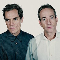 Two men stand close together against a plain light background, one in a dark striped shirt and the other in a white shirt, both wearing serious expressions, creating a somber and introspective mood.
