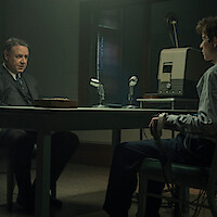 Two men sit across a table in a dimly lit interrogation room, one wearing a suit and the other connected to a polygraph machine, microphones and recording equipment between them.