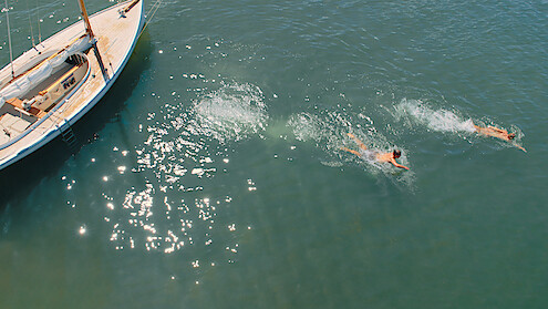 A man and a woman swim away from a sailing boat in a beautiful, sun-lit sea.