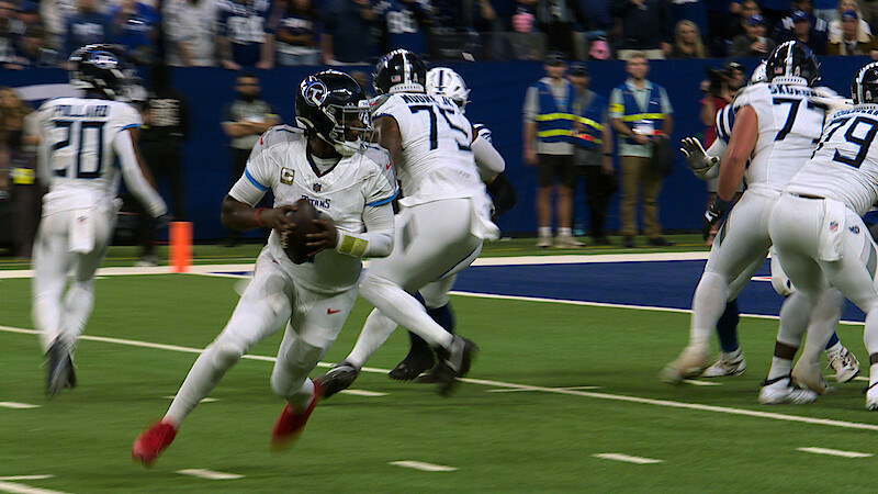 Football quarterback in white uniform running with the ball on a green field during a game, with teammates blocking and stadium spectators in the background.