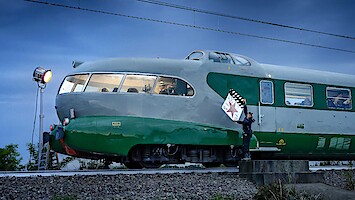 A vintage green and gray streamlined train is stopped outdoors at dusk on train tracks; a person in uniform stands beside it holding a clapperboard while a film light illuminates the scene, creating a cinematic atmosphere.