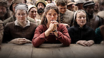 A distressed young woman in historical clothing sits at a wooden table, hands clasped, surrounded by somber villagers in period attire, suggesting a dramatic or tense moment in a historical community setting.