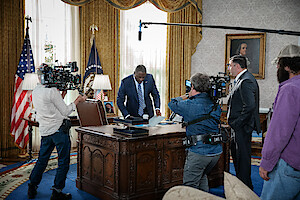 Actor in a suit stands at a presidential desk in an Oval Office set, surrounded by film crew, cameras, and lighting equipment, creating a busy, professional atmosphere for a political drama scene.