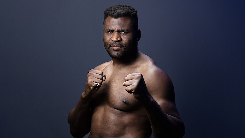 Francis Zavier Ngannou in a fighting stance with clenched fists, looking serious, posing against a plain dark gradient background in a studio setting.