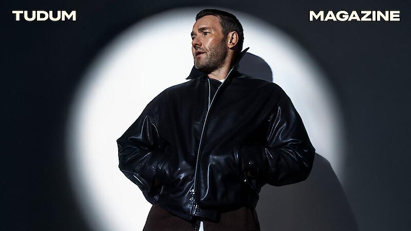 Joel Edgerton illuminated by a spotlight in front of a white wall wearing a black leather jacket looking to the side. 