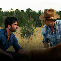 Two men kneel in a grassy field framed by trees. Yancy Grey (Jack Schumacher) wears a blue denim shirt, while Staten Kirkland (Josh Duhamel) sports a denim shirt and a cowboy hat.