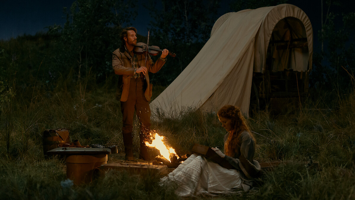 Man playing violin by campfire while woman reads beside him, set in grassy outdoors at night with a covered wagon and camping supplies in the background, evoking a historical or pioneer setting.