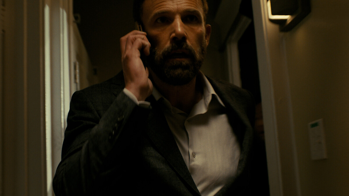 A man in a dark suit talking on a phone walks through a dimly lit hallway, looking serious and focused, with warm lighting on the walls and a tense atmosphere.