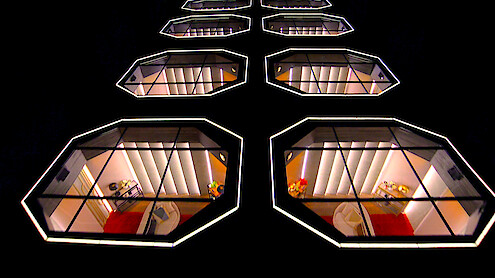 Rows of illuminated, modern geometric windows on a dark building facade at night, each showing stylish interiors with red and orange accents, chairs, and small tables, creating a futuristic architectural pattern.