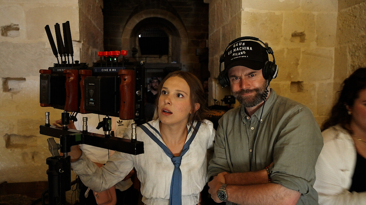 A woman in costume and a man with headphones stand by a camera rig in a stone hallway on a film set, reviewing footage together.