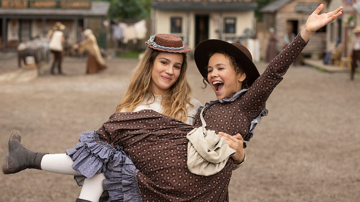 Two women in period costumes smile joyfully in an old western town setting; one lifts the other while she raises her arm in excitement, with rustic buildings and people in the background.