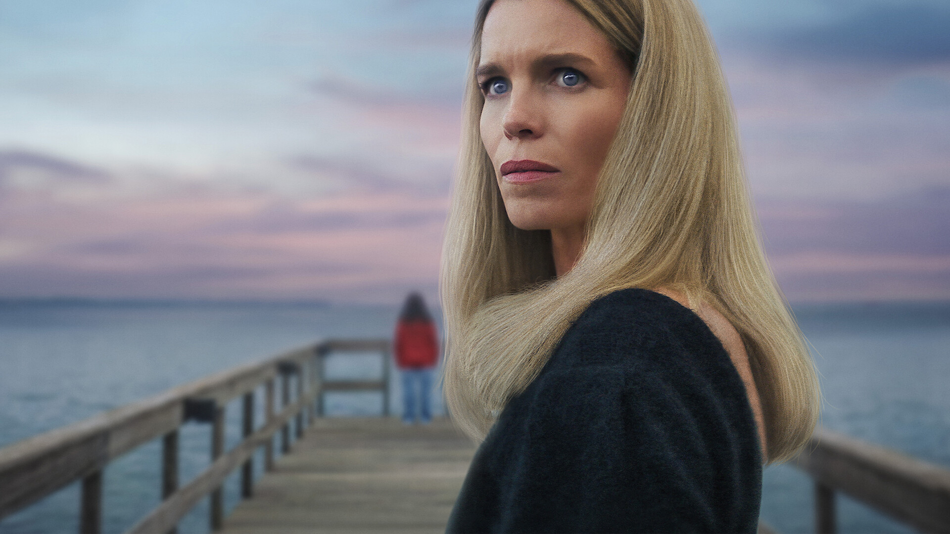 Secrets We Keep - A woman looking pensive standing on a dock while a mysterious person in red stands in the background