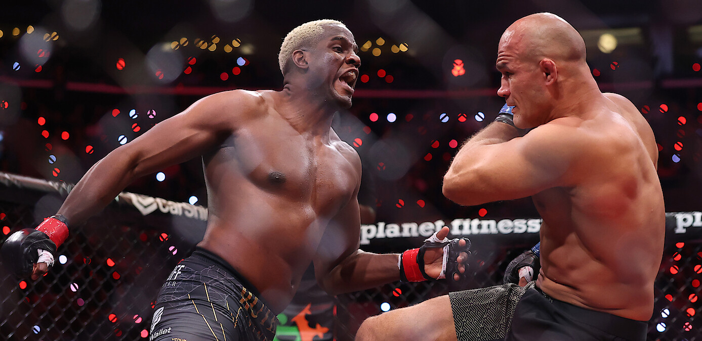 Two male MMA fighters compete intensely inside a caged octagon during a professional match, with one throwing a punch and the other defending himself. Arena lights and a crowd are visible in the background.