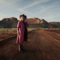 A woman in a pink coat and cowboy hat stands on a path in a Southwestern landscape.
