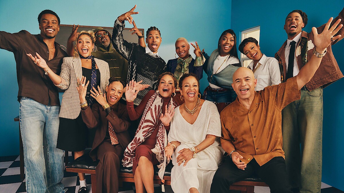 Group of people in stylish clothing smiling and posing energetically in a brightly lit room with blue walls and checkered floor, conveying a sense of joy and celebration.