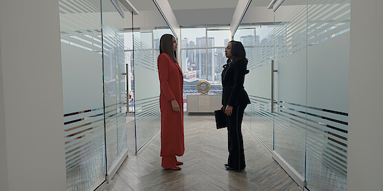 Crystle Stewart as Mallory and Taylor Polidore Williams as Kimmie in business attire stand facing each other in a modern office hallway with glass walls, city skyline visible through large windows in the background.