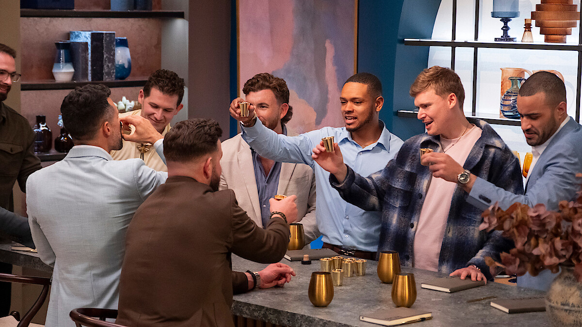 A group of men standing and sitting around a modern kitchen island, raising drinks in a toast. Shelves and decorative objects are visible in the background, creating a warm and social atmosphere.