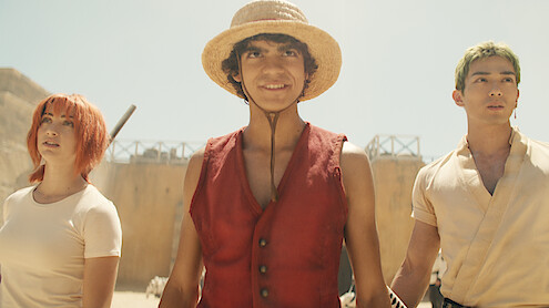 Three young people stand outdoors under bright sunlight in a sandy, desert-like environment. The central character wears a straw hat and red vest, flanked by companions with distinct hair colors and casual clothing, looking determined.