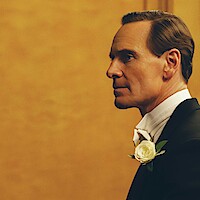 Man in formal attire with a white bow tie and flower boutonniere stands in profile against a plain golden background, looking thoughtful and composed in a well-lit indoor setting.