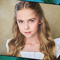 Young girl with long wavy blonde hair wearing a white blouse, sitting indoors against a neutral beige background, looking directly at the camera with a calm expression.
