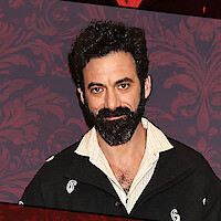 Man with curly dark hair and beard standing indoors against a decorative purple patterned wall, wearing a black jacket over a light-colored shirt, facing the camera and smiling softly.