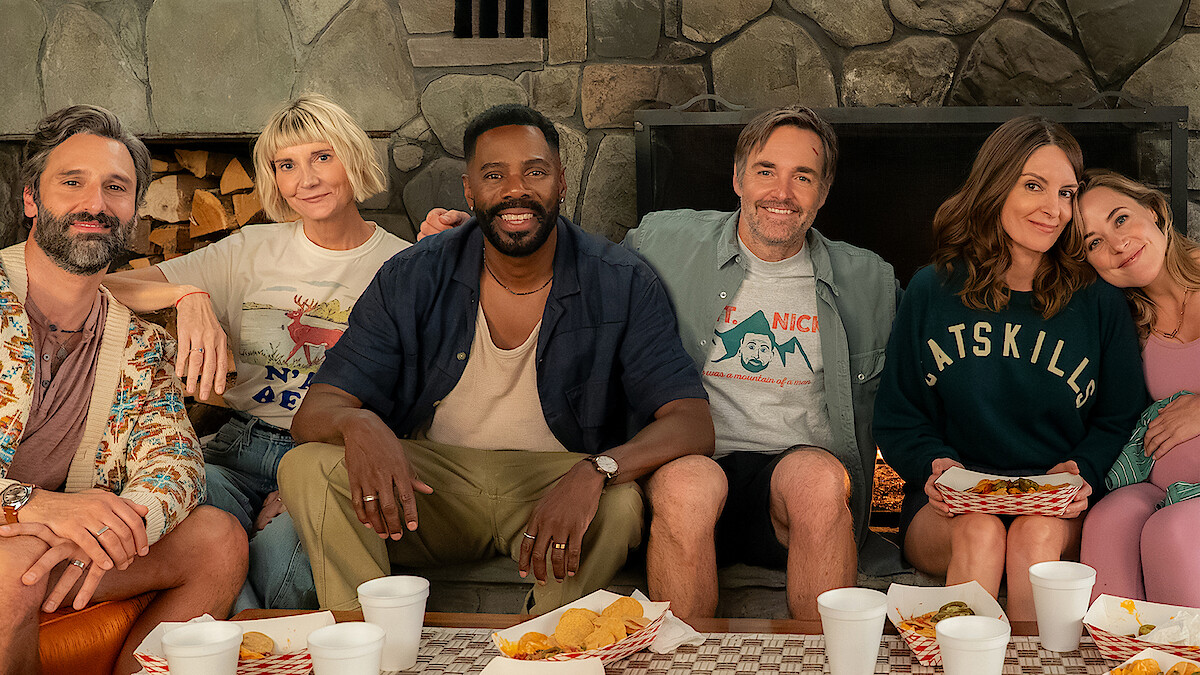 Six adults sitting close together on a couch in a cozy room with a stone fireplace, enjoying a casual meal with trays of food and drinks, smiling and relaxed in a friendly, informal setting.