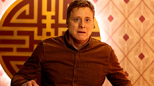 A man in a brown shirt looks surprised in a warmly lit room with patterned wallpaper and a large decorative circular wall design behind him.