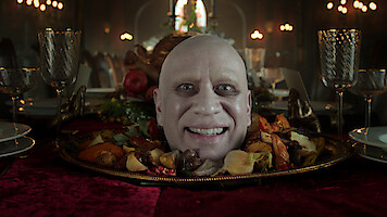 A smiling bald man’s head appears on a platter surrounded by roasted vegetables, set on an elegant dining table with ornate glasses and a dimly lit, candlelit gothic banquet hall in the background.