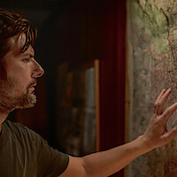 Man closely examining a large map dotted with red pins in a dimly lit room, appearing focused and thoughtful as he touches the map with his hand.