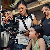 A woman shows a young girl and a group of people how to use a camera on a tripod in a market setting. The mood is focused and friendly, suggesting collaboration and learning in a lively outdoor environment.