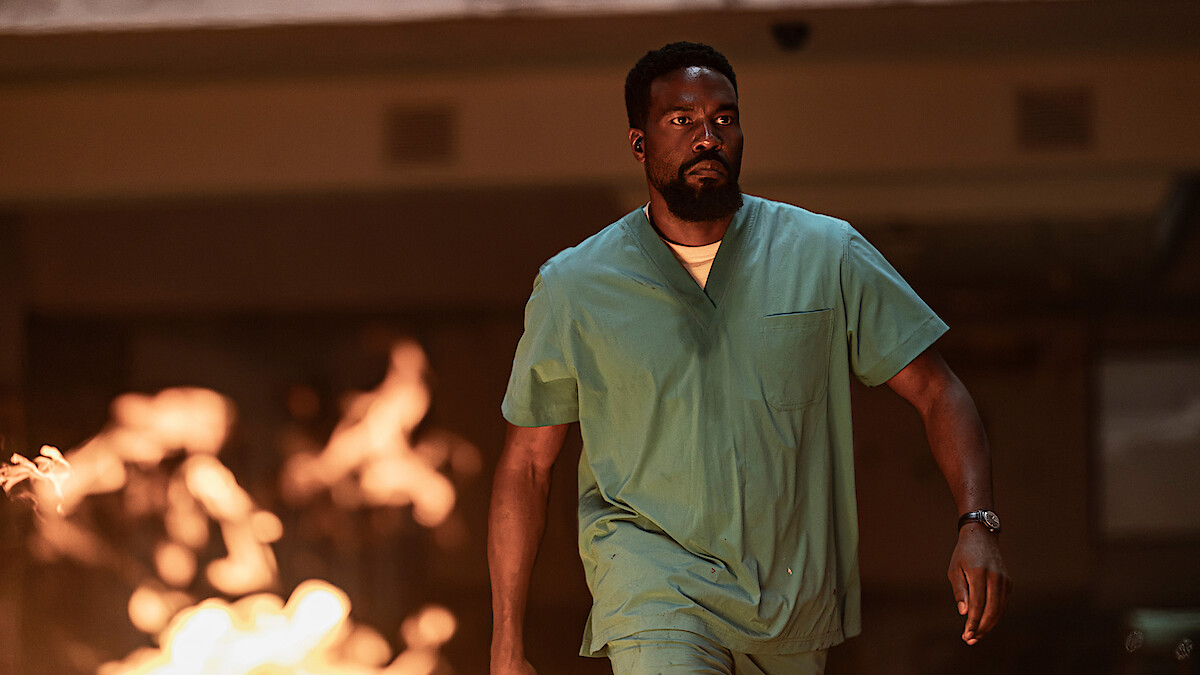 Man in green scrubs walking with determination through a smoky indoor environment with flames in the foreground, suggesting urgency or danger.