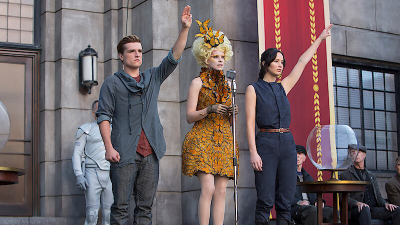 Three people stand in front of a stone building, two raising one hand; one wears a grey shirt, another a sleeveless outfit, and the third is in an elaborate orange dress at a microphone. Others are seated nearby, and a large banner hangs.