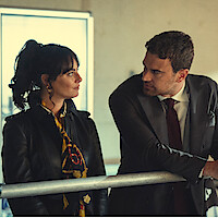 A woman in a leather jacket and scarf stands next to a man in a suit and tie. They are indoors, leaning on a metal railing, looking at each other in a serious conversation in a modern, softly lit room.
